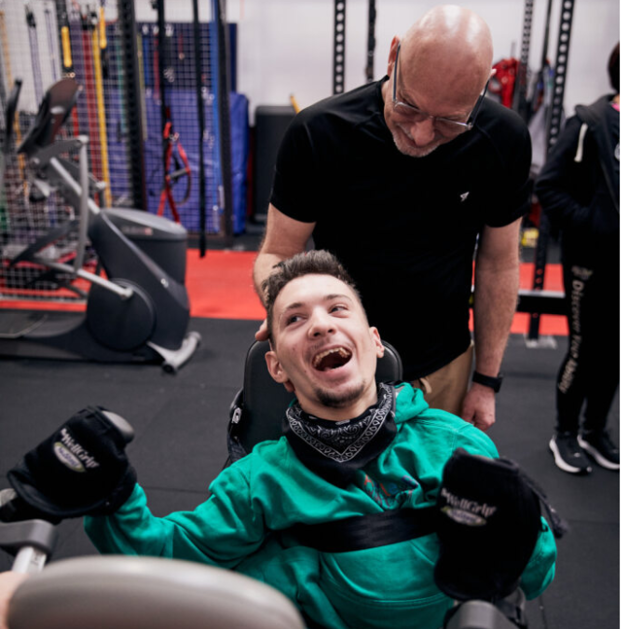 The photo is taken inside the Able2B gym. A young man with brown short hair, navy scarf and green jumper is using an exercise machine smiling. Behind the young man is a man in a black top and brown joggers. He is holding the young man’s head to ensure he is sitting correctly when using the exercise machine. Those photographed are Warren and Tyler.