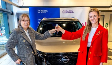 The picture has been taken in Bristol Street Motors Nissan, Halifax. Kate Dearden MP is presenting car keys to Allison. There is a silver car parked behind them.
