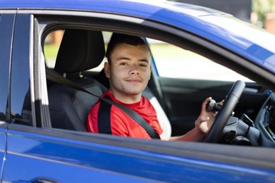 A young man is sitting and smiling in the front driving seat of his blue vehicle. He is wearing a red top with brown short hair.