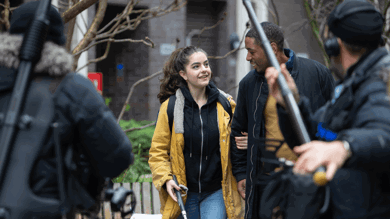 The image is of behind the scenes filming of Nyla's Journey film. There is a man featured in the foreground with a boom mic pointed at Nyla and her helpful companion walking down the road. Nyla is using a walking cane.