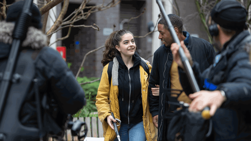 The image is of behind the scenes filming of Nyla's Journey film. There is a man featured in the foreground with a boom mic pointed at Nyla and her helpful companion walking down the road. Nyla is using a walking cane.