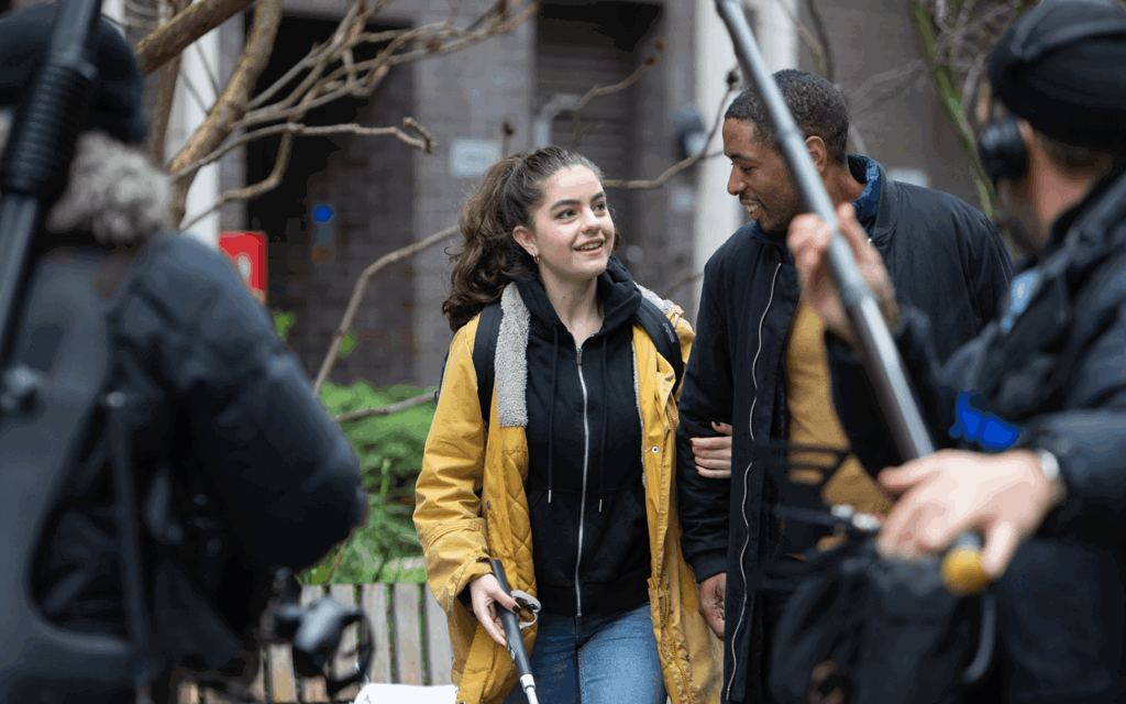 The image is of behind the scenes filming of Nyla's Journey film. There is a man featured in the foreground with a boom mic pointed at Nyla and her helpful companion walking down the road. Nyla is using a walking cane.