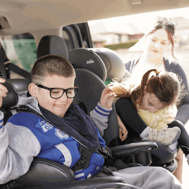 Gino is sat in his wheelchair in his WAV, next to him is his mum who is plugging his sister into a car seat.