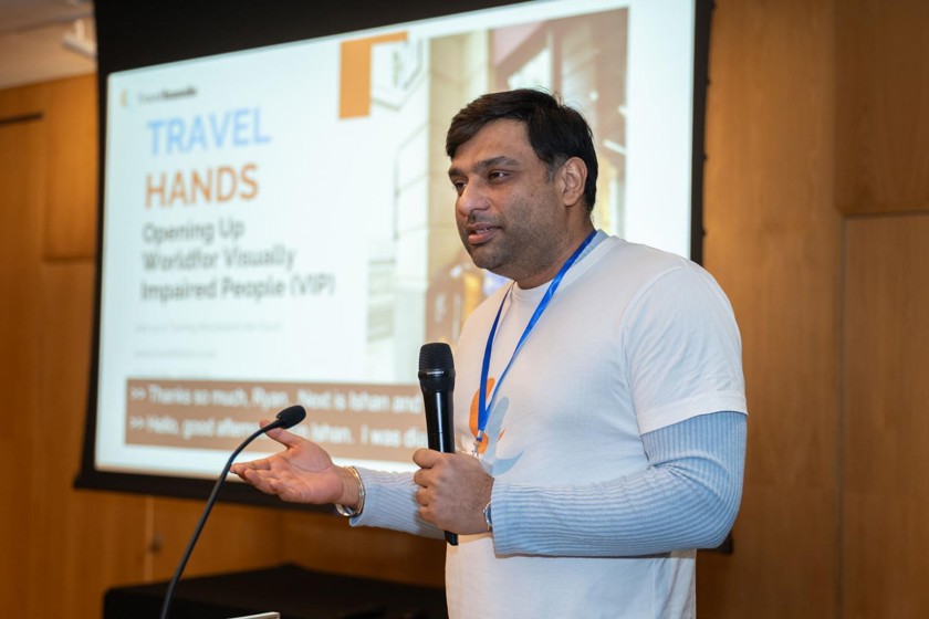 A man is presenting with a mic in his hand at a lectern. He is wearing a white and blue striped long sleeve top with a white t-shirt over the top with the Travel Hands logo. 