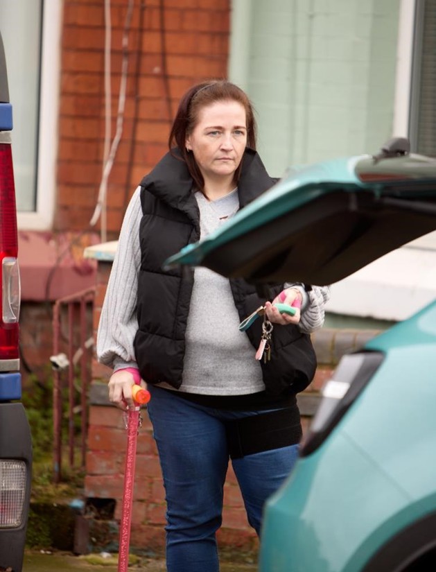 Leanne is closing the boot of her blue vehicle using her car key. She has long brown hair, black gilet, grey jumper, blue jeans and a orange walking aid.