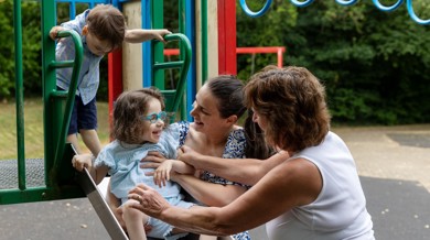 Boronica is sat on a slide smiling. She has long brown curly hair, blue rimmed glasses and a white and blue top and shorts on. Boronica’s mum Crystal, is on her right holding her and the slide and Boronica’s grandmother is doing the same.