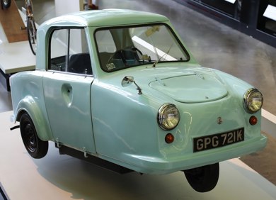 A parked three wheeled blue trike car within a building. 