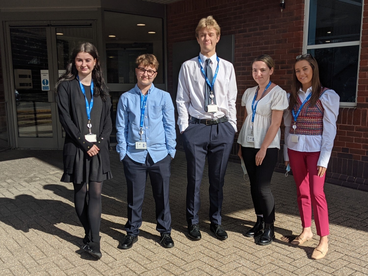 A picture of two young men and three young women standing outside a building smiling. 