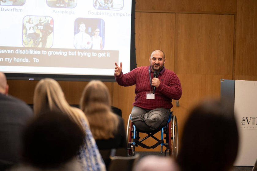 A man is presenting on stage to an audience. He is a wheelchair use. The man is wearing a read jumper and grey jeans. 