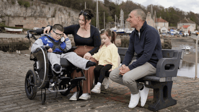 Gino is sat in his wheelchair next to his mum, sister and dad, who are sat on a bench. They are laughing and talking. Behind them are boats and cottages next to the sea. 