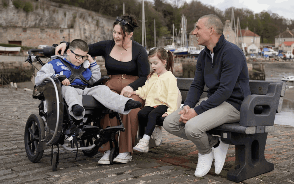 Gino is sat in his wheelchair next to his mum, sister and dad, who are sat on a bench. They are laughing and talking. Behind them are boats and cottages next to the sea. 