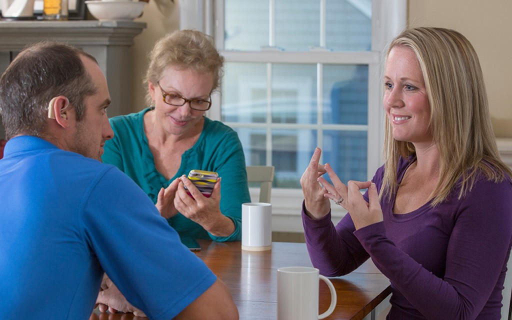 A man and a women sat at a wooden table with a British Sign Language interpreter. 