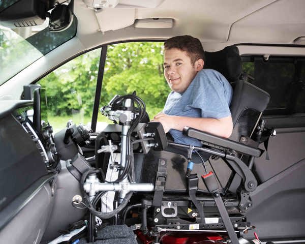 A boy is in a wheelchair smiling in the front of a car. 