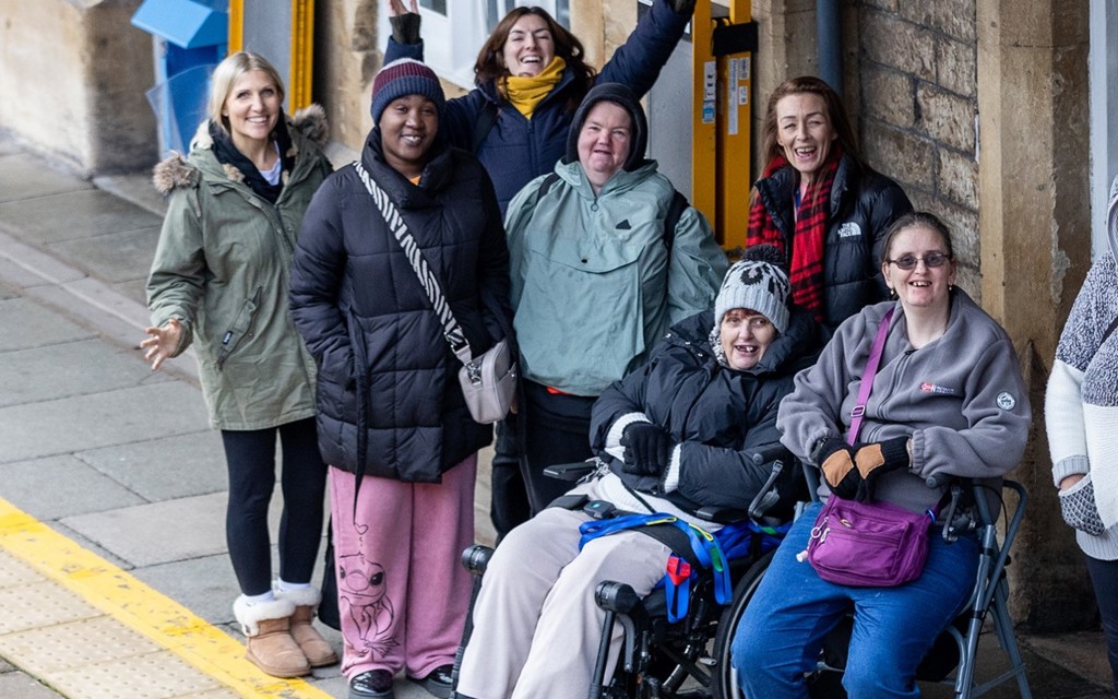 A group of women are laughing and posing for the photograph which is taken at a distance. They are standing or sitting on a train platform.