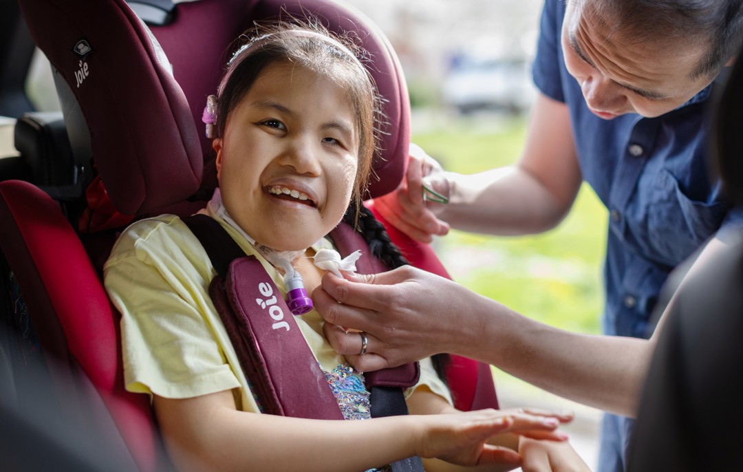 Eva is smiling at the camera in a yellow top, pink headband, she is sitting in her bright pink car seat. Eva's dad is helping her into the car seat smiling. He is in a navy blue button up short sleeved shirt.