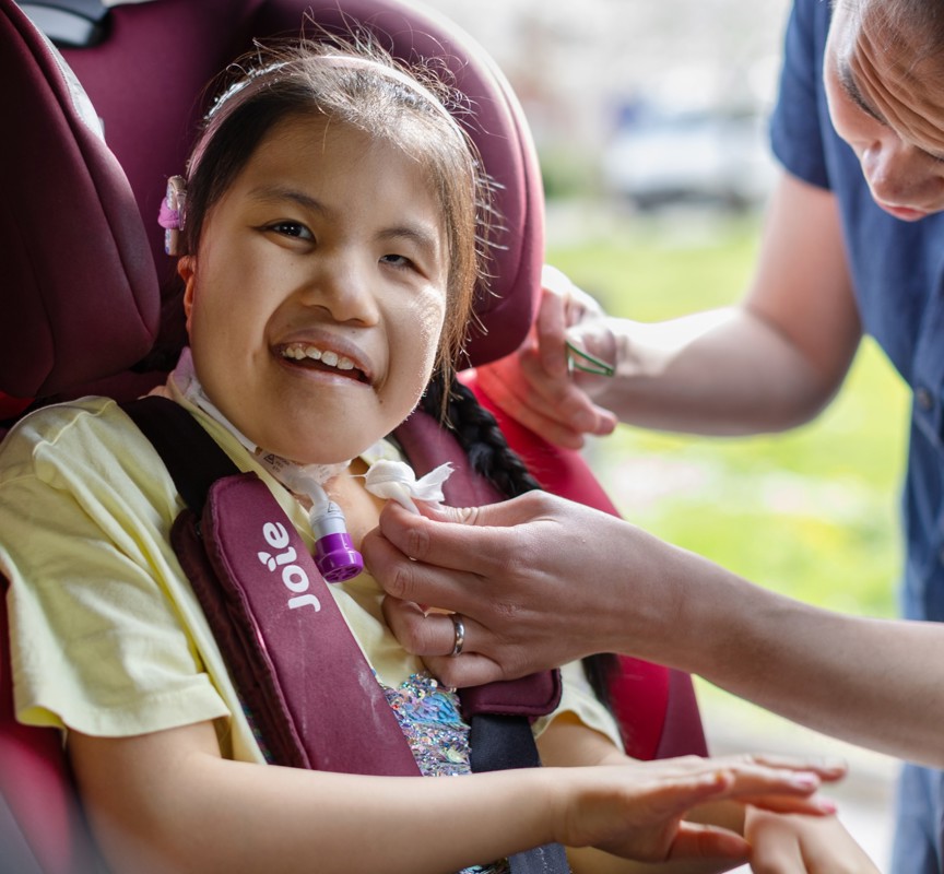 Eva is smiling at the camera in a yellow top, pink headband, she is sitting in her bright pink car seat. Eva's dad is helping her into the car seat smiling. He is in a navy blue button up short sleeved shirt.