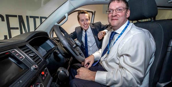 The photo shows the inside of an adapted vehicle. Sat in the front seat of the vehicle is a Motability employee smiling with his hand on knees, wearing a smart suit. Tom Pursglove MP is smiling and looking in the vehicle from outside next to the employee with his arm rested on the open right front door.