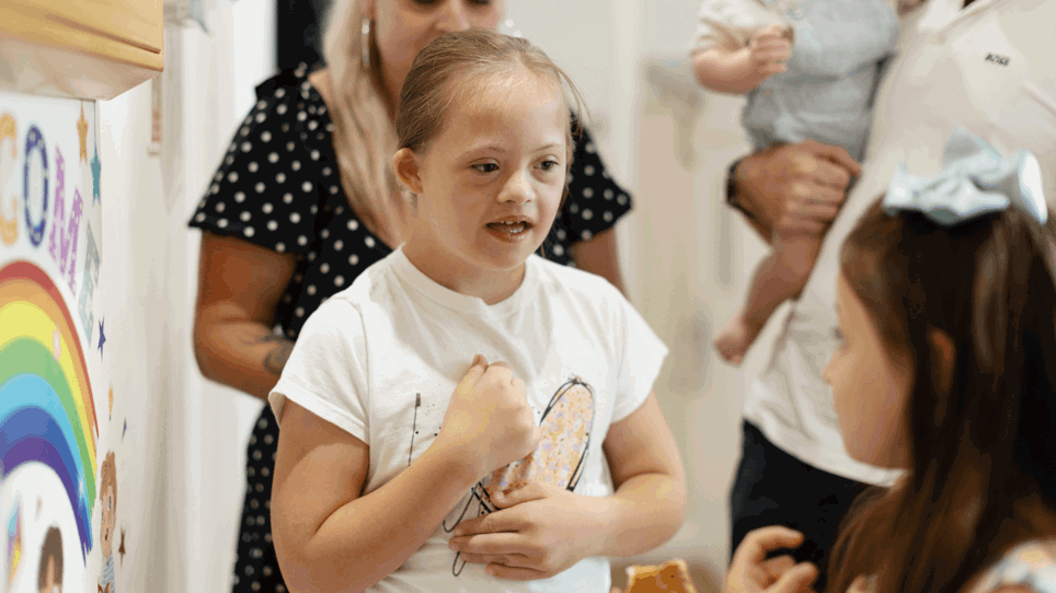 Macie is standing and looking at her sister. Macie is in a white t-shirt with a large pink heart in the centre. She has long plaited brown hair.