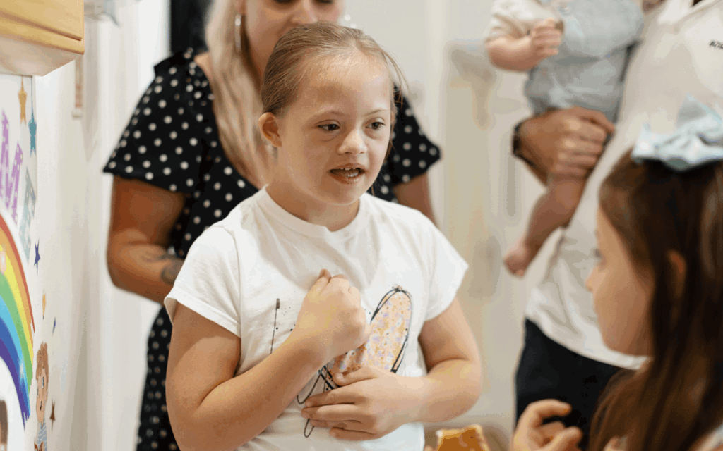 Macie is standing and looking at her sister. Macie is in a white t-shirt with a large pink heart in the centre. She has long plaited brown hair.
