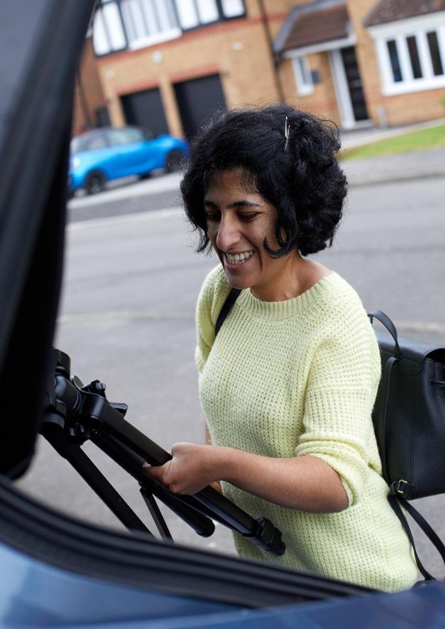 Mathy is smiling while taking her black camera tripod out of the boot of her blue vehicle. She is wearing a yellow knitted jumper, black backpack and has short black hair in a bob.