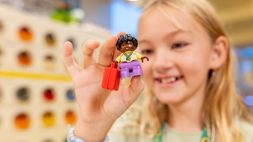 A young girl with long blonde hair and a green jumper wearing a hidden disability sunflower lanyard. She is holding a Lego minifigure in her fingers with a hidden disability sunflower lanyard.