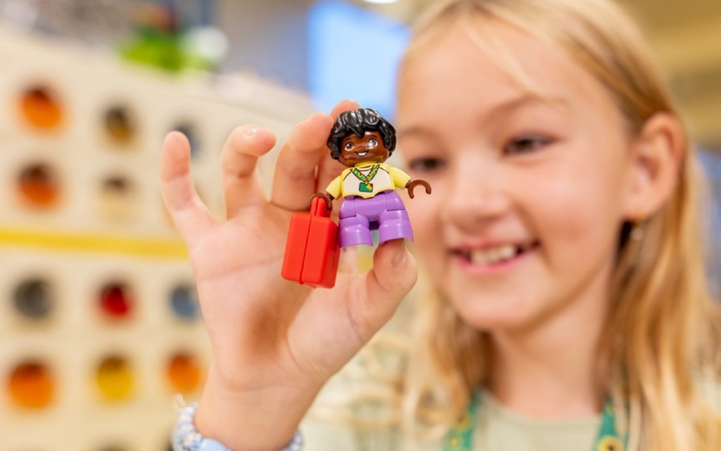 A young girl with long blonde hair and a green jumper wearing a hidden disability sunflower lanyard. She is holding a Lego minifigure in her fingers with a hidden disability sunflower lanyard.