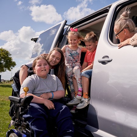 A boy sits in a powered wheelchair alongside his family next to their wheelchair accessible vehicle.