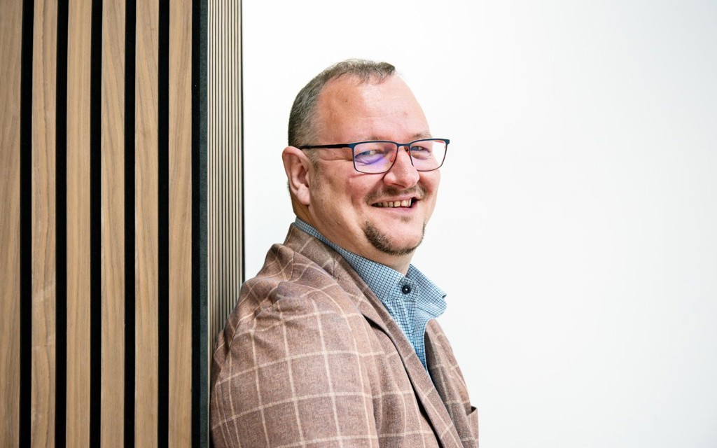 Nick is smiling, he is wearing black rimmed glasses, blue shirt and brown jacket.
