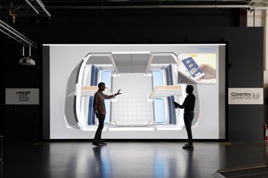 Two men are standing in a room and chatting. A large screen is behind them.