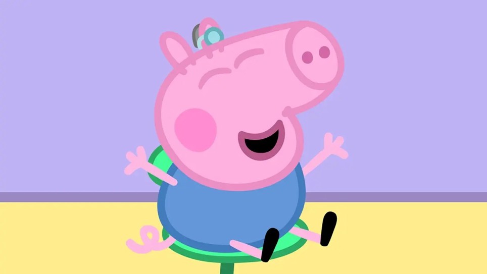 George from Peppa Pig is sitting on a seat with a hearing aid in his right ear. He has a big smile on his face and his arms are out stretched. 