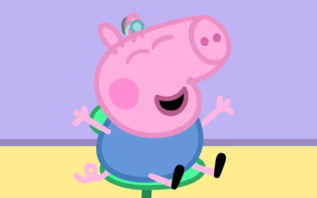 George from Peppa Pig is sitting on a seat with a hearing aid in his right ear. He has a big smile on his face and his arms are out stretched. 