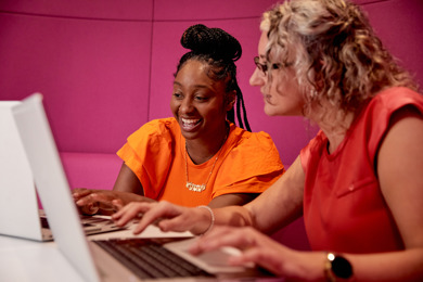 Two women sit at a table looking at a computer