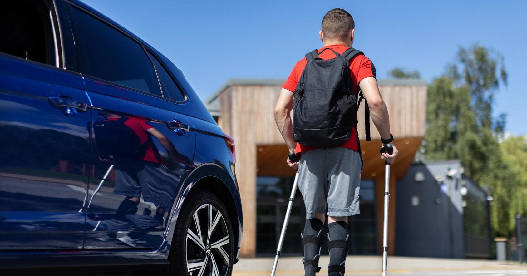 A young man is walking away from his blue vehicles using crutches. He had a red top on with a black backpack and blue shorts. 