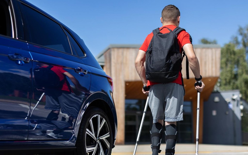 A young man is walking away from his blue vehicles using crutches. He had a red top on with a black backpack and blue shorts.