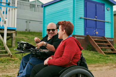 Angela and Pete and both laughing and talking whilst wheeling down the sea front. Behind them are brightly painted beach huts. 