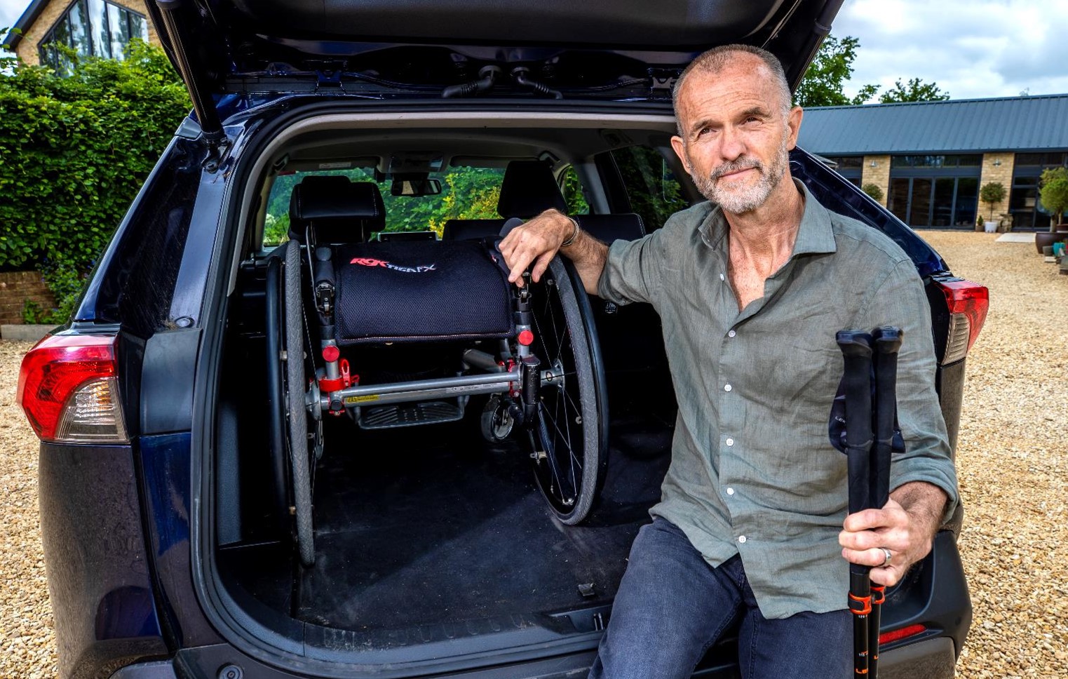 Paul is sitting in the boot of his black car with his arm on his folded up black wheelchair. Paul is smiling. Paul has grey short hair, wearing a grey button up shirt and blue jeans. 