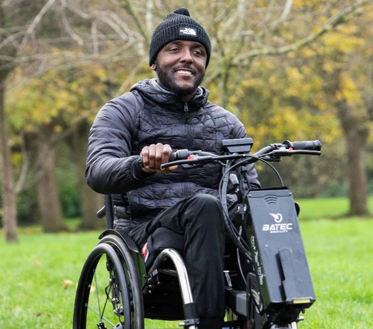 Image of a gentleman smiling while he wheels in a powered wheelchair. There a big green trees behind him. 