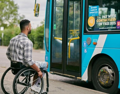 A man with short brown hair is sitting in a wheelchair on the side of the road ready to wheel into a parked blue bus. 