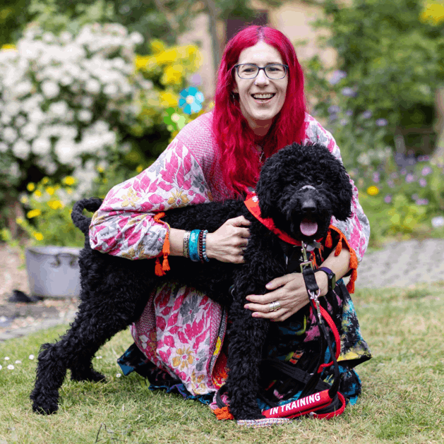 Charlie has pink hair and is wearing multi coloured clothing. They are holding their dog by the lead in their garden. Their dog is black poodle cross and is wearing a red neckerchief with a "in training" sign on their lead.