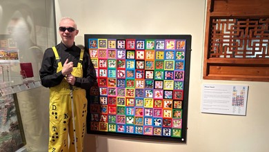 Mr Dot is photographed in front of an art piece. He is wearing black glasses, a black shirt and yellow dungarees.