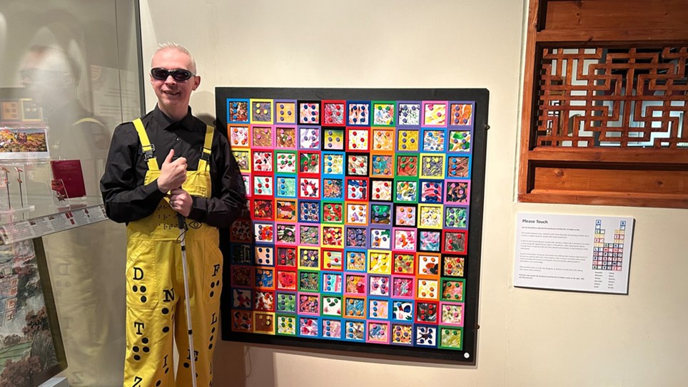Mr Dot is photographed in front of an art piece. He is wearing black glasses, a black shirt and yellow dungarees.