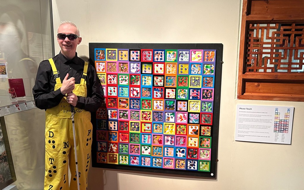 Mr Dot is photographed in front of an art piece. He is wearing black glasses, a black shirt and yellow dungarees.