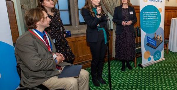 A lady is standing and talking into a microphone whilst two ladies and a man in a wheelchair listen.