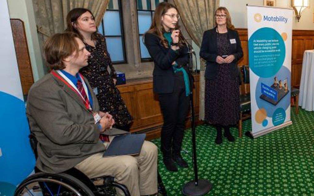 A lady is standing and talking into a microphone whilst two ladies and a man in a wheelchair listen.
