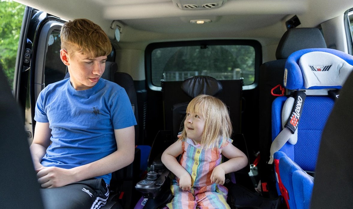 Poppy is sitting in her powered wheelchair in her vehicle. To the left of her is her old brother Liam, Poppy and Liam and smiling at one another. To the right of her is her car seat.