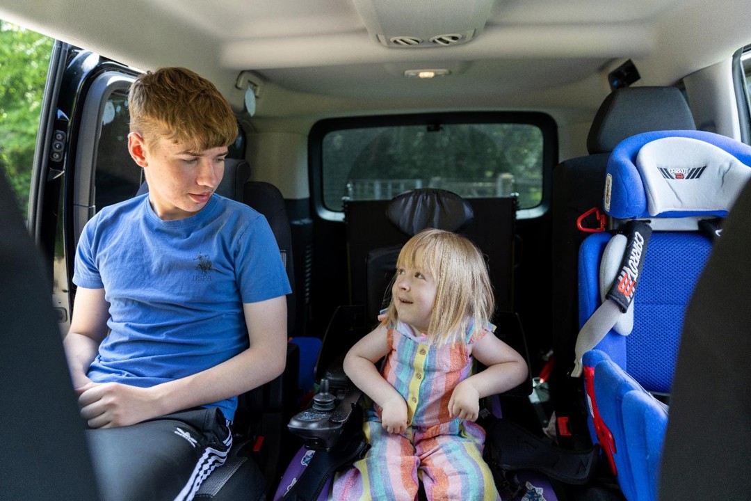 Poppy is sitting in her powered wheelchair in her vehicle. To the left of her is her old brother Liam, Poppy and Liam and smiling at one another. To the right of her is her car seat.
