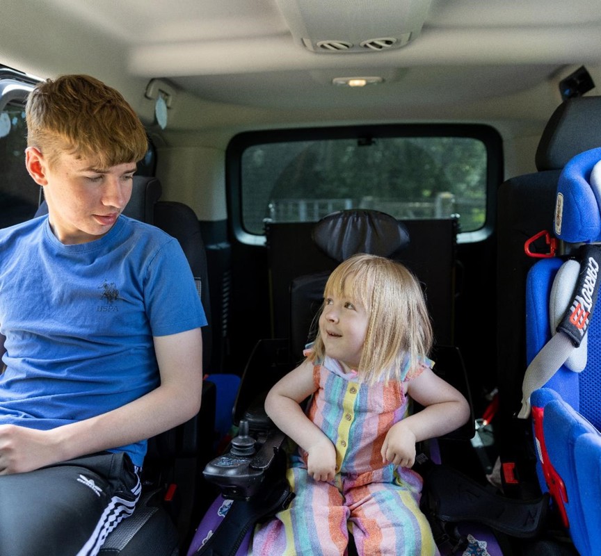 Poppy is sitting in her powered wheelchair in her vehicle. To the left of her is her old brother Liam, Poppy and Liam and smiling at one another. To the right of her is her car seat.