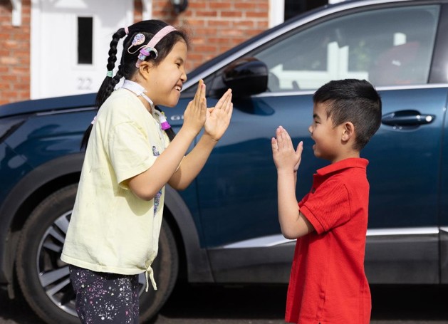 Eva is playing clapping games in a yellow top, pink headband, night sky patterned trousers. Standing in front of her playing clapping games with her is her brother in a red polo top. They are both laughing.
