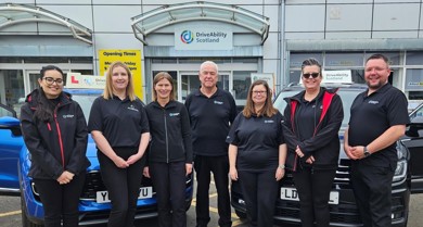 A group photo of five women and two men standing in front of two Driving Mobility Scotland cars smiling in black polos, black trousers and black trainers.