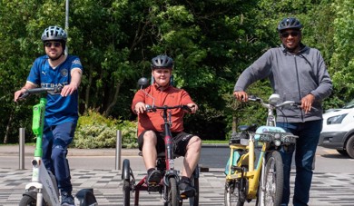 The image is of three men using micromobility equipment, one e-scooter, one cycle and one e-bike, outside in a park. The sun is shining.